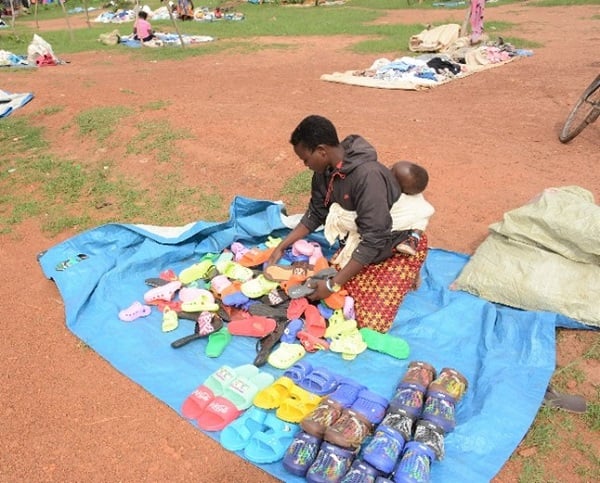 Girl sits on blanket with baby on her back at a market in Uganda, surrounded by shoes.