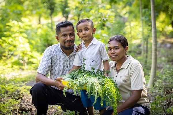 A mom, dad and little boy in Indonesia stand together with vegetables from their garden, smiling.