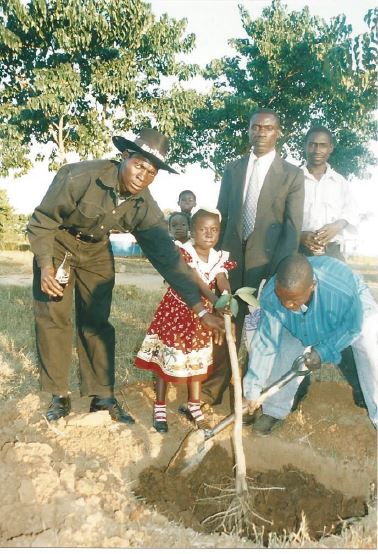 A young girl in Zambia plants a tree, surrounded by community members.