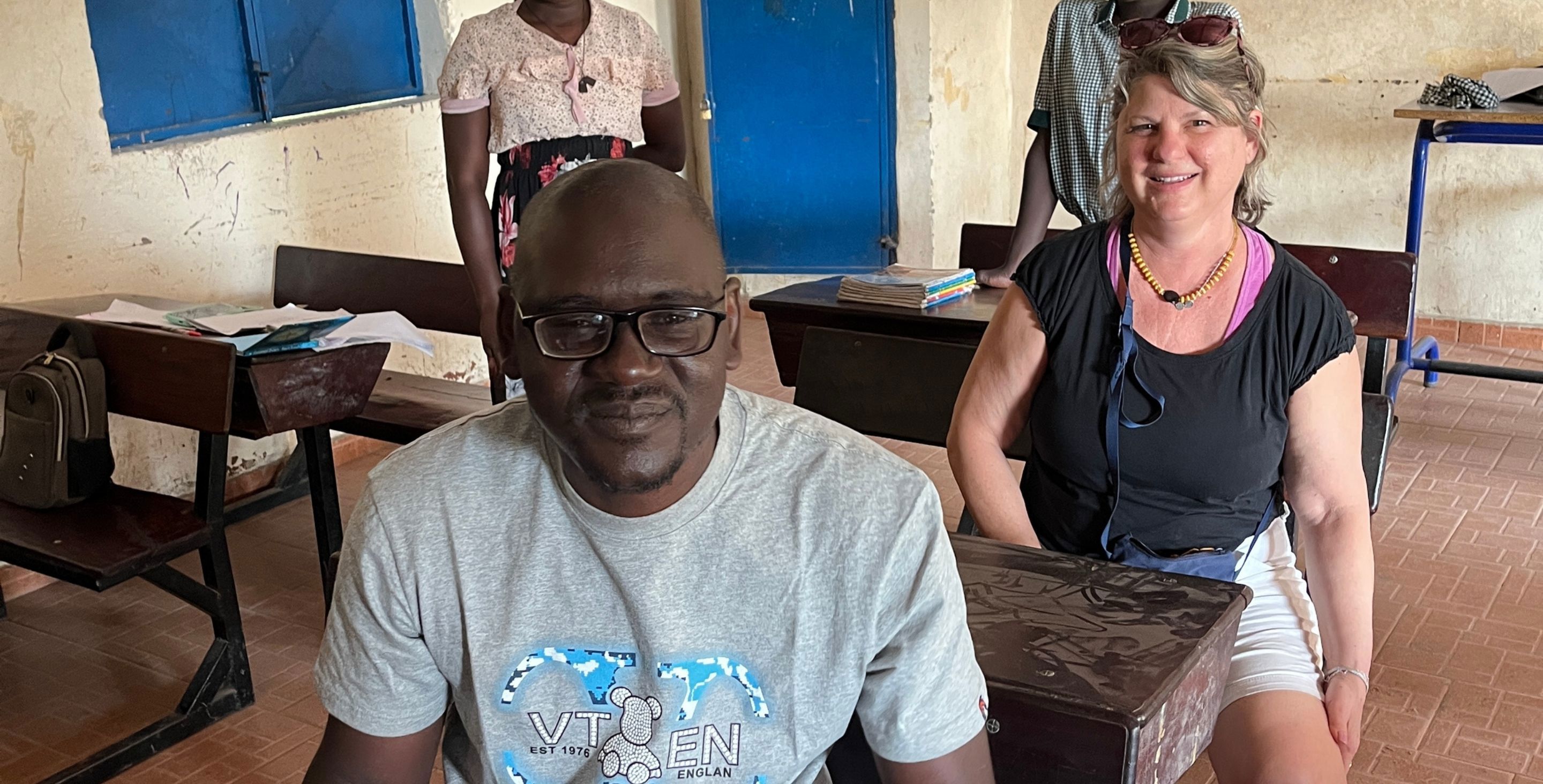 An African man and American woman sit at small desks in a classroom in The Gambia, smiling at the camera.