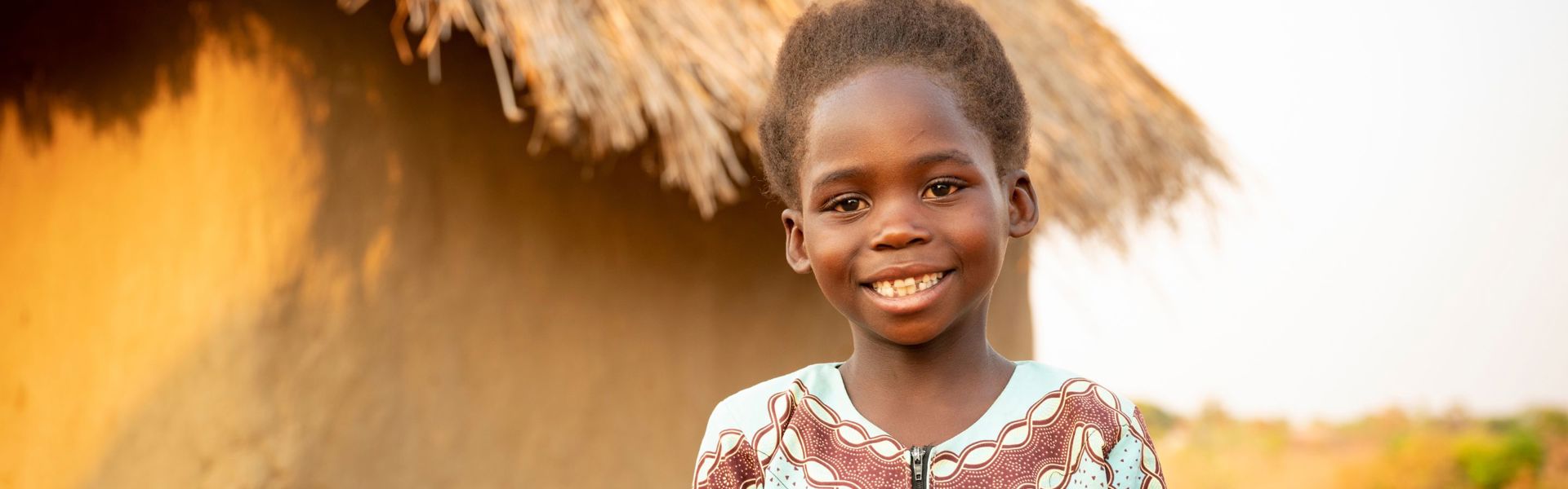 A girl in Zambia wearing traditional clothing stands outside smiling.