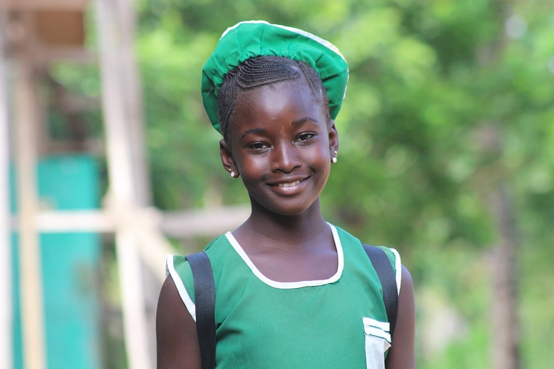 Schoolgirl in green uniform stands smiling in front of her school in Sierra Leone.