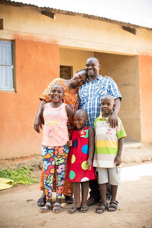 Middle aged man in Uganda stands smiling with his wife and three children.