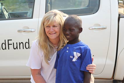 Sponsor and sponsored child stand outdoors in Zambia, hugging and smiling.