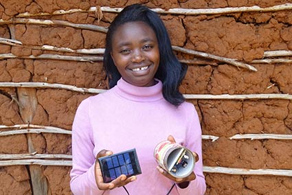 Teenager in pink shirt stands in front of mud wall in Kenya, holding solar-powered light, smiling.
