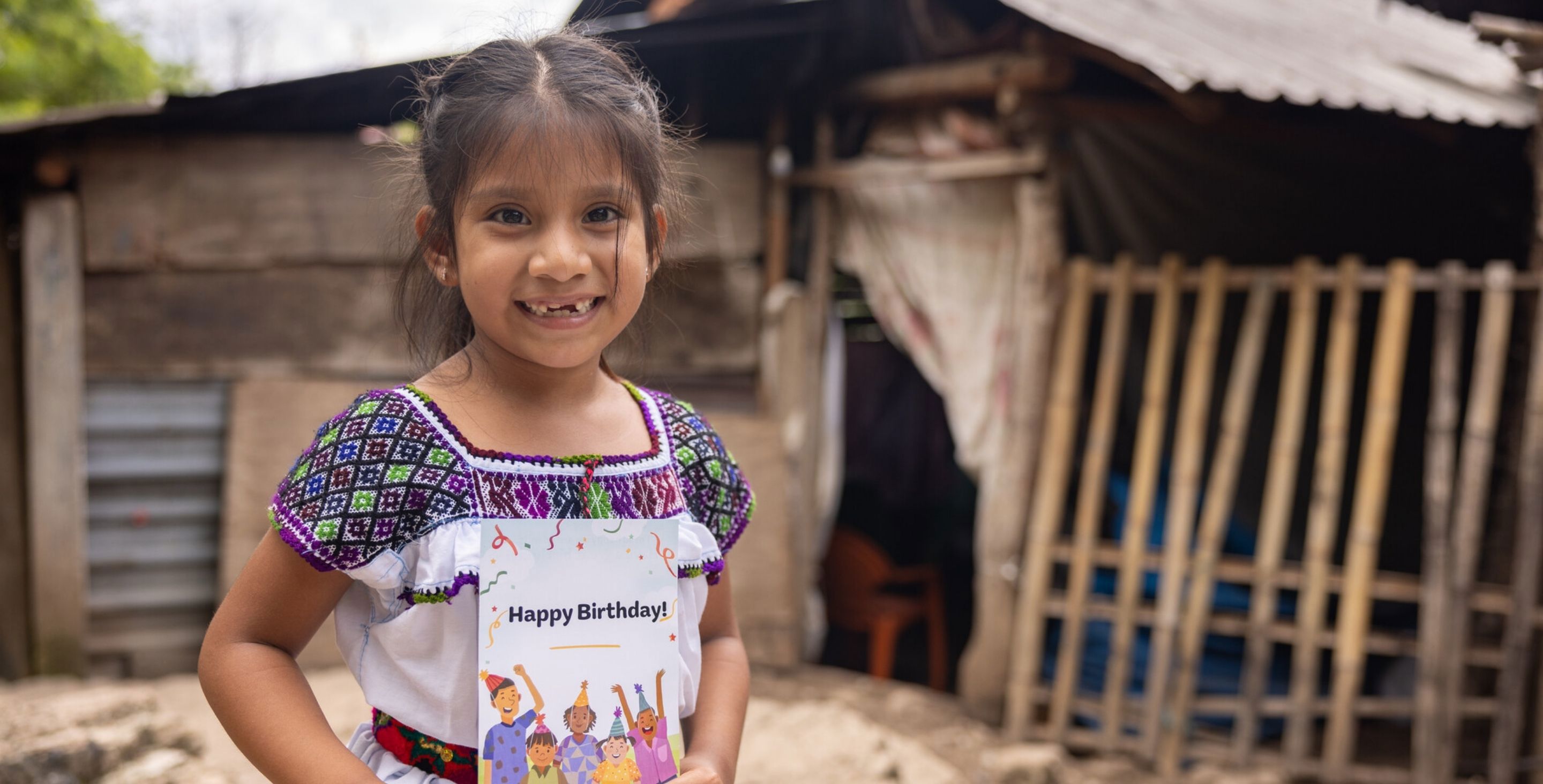 A little girl sits outside her home in rural Mexico, holding a card that says 