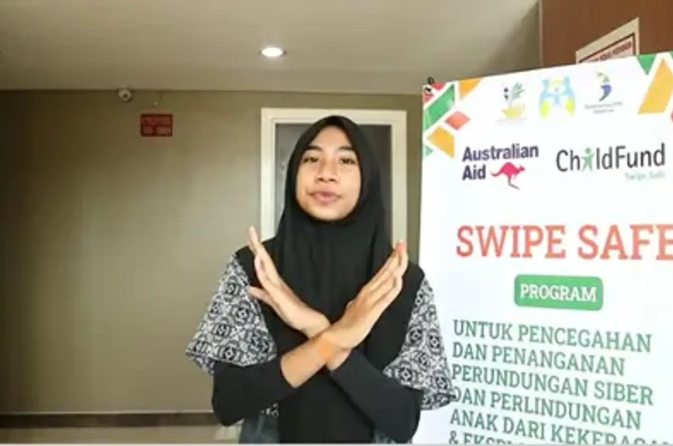 A person makes an “X” gesture with their arms in front of a Swipe Safe program banner promoting digital literacy and online child protection, part of a ChildFund and Australian Aid initiative.