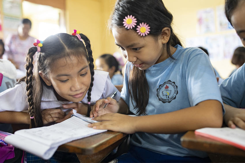 Two girls sit at a desk at school in the Philippines, writing.
