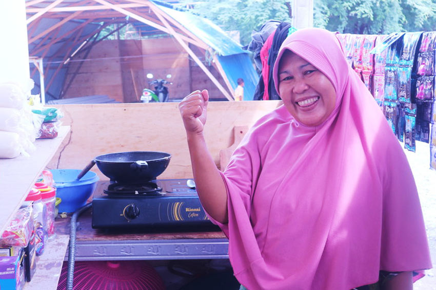 A woman in pink hijab stands inside a snack shop by a cooking pot, flexing her arm and smiling.