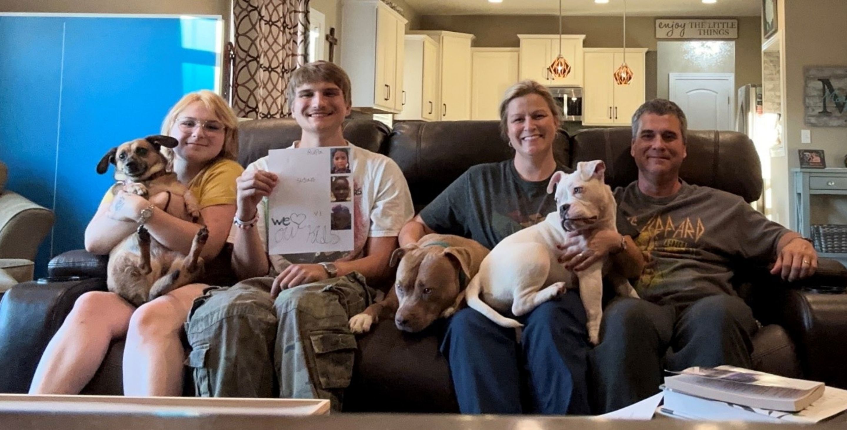 A family consisting of a mom, dad, two teenage kids and a dog sit on a couch in the U.S., smiling at the camera. They are holding up a piece of paper showing photos of their sponsored children.