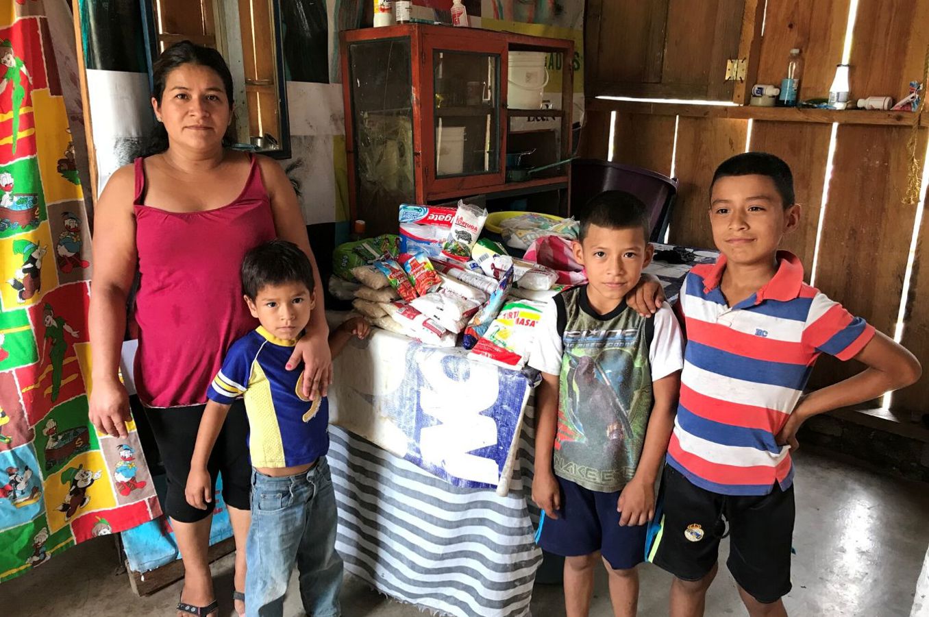 A mother and her three sons stand around a table in a modest, wooden house in Honduras. On the table are food items.