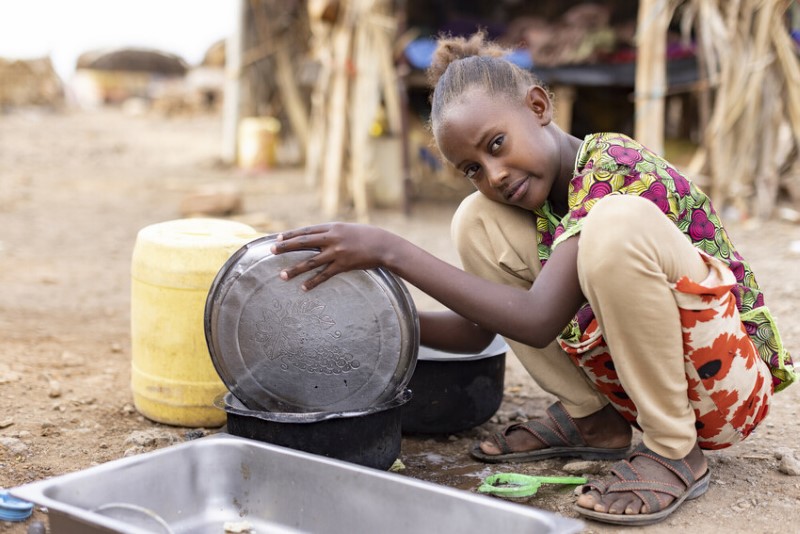 A teen girl in Kenya washes dishes while looking somberly at the camera.