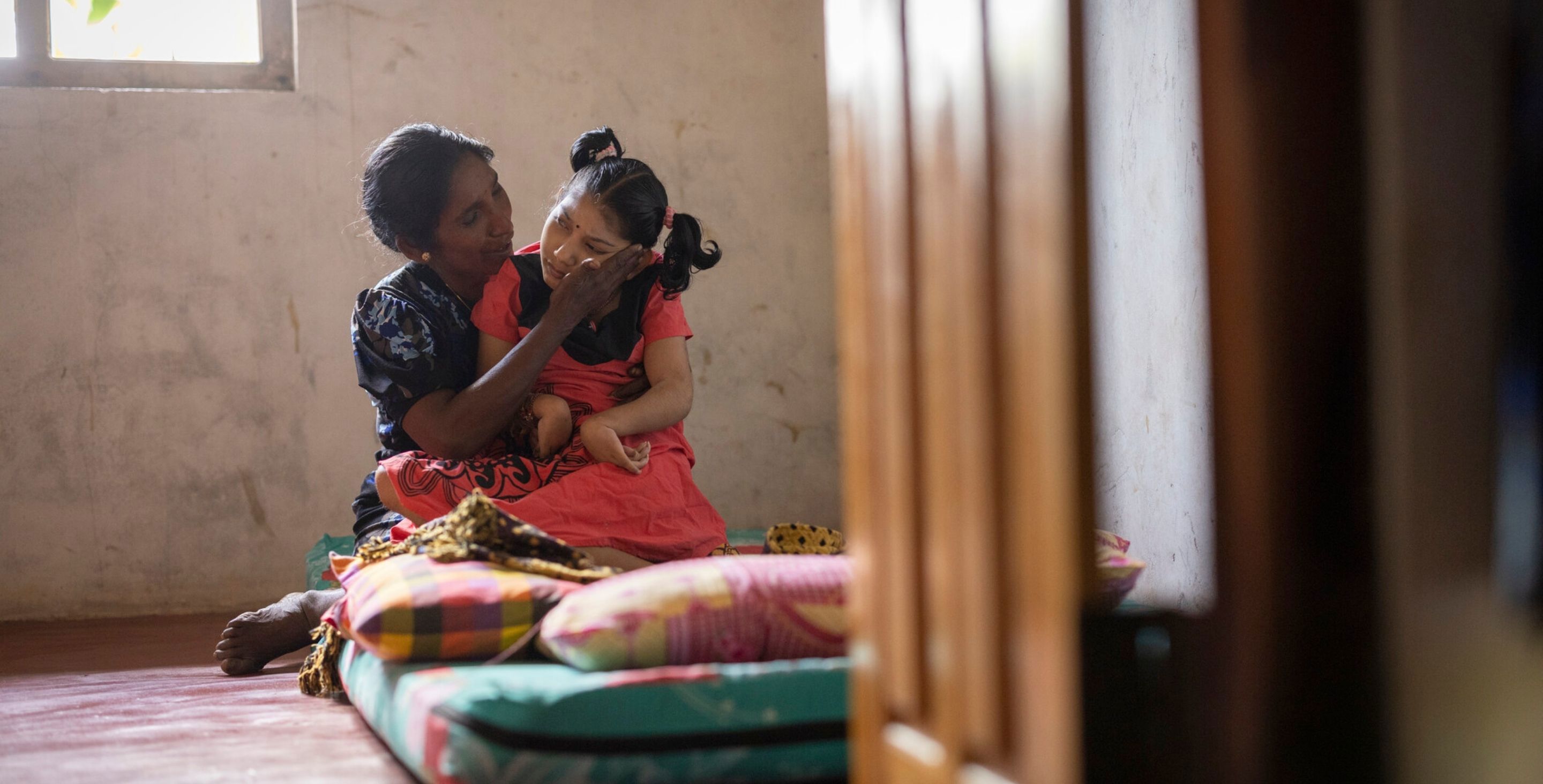 A mother sits on a mat with her daughter, who has cerebral palsy, on the floor of their home.