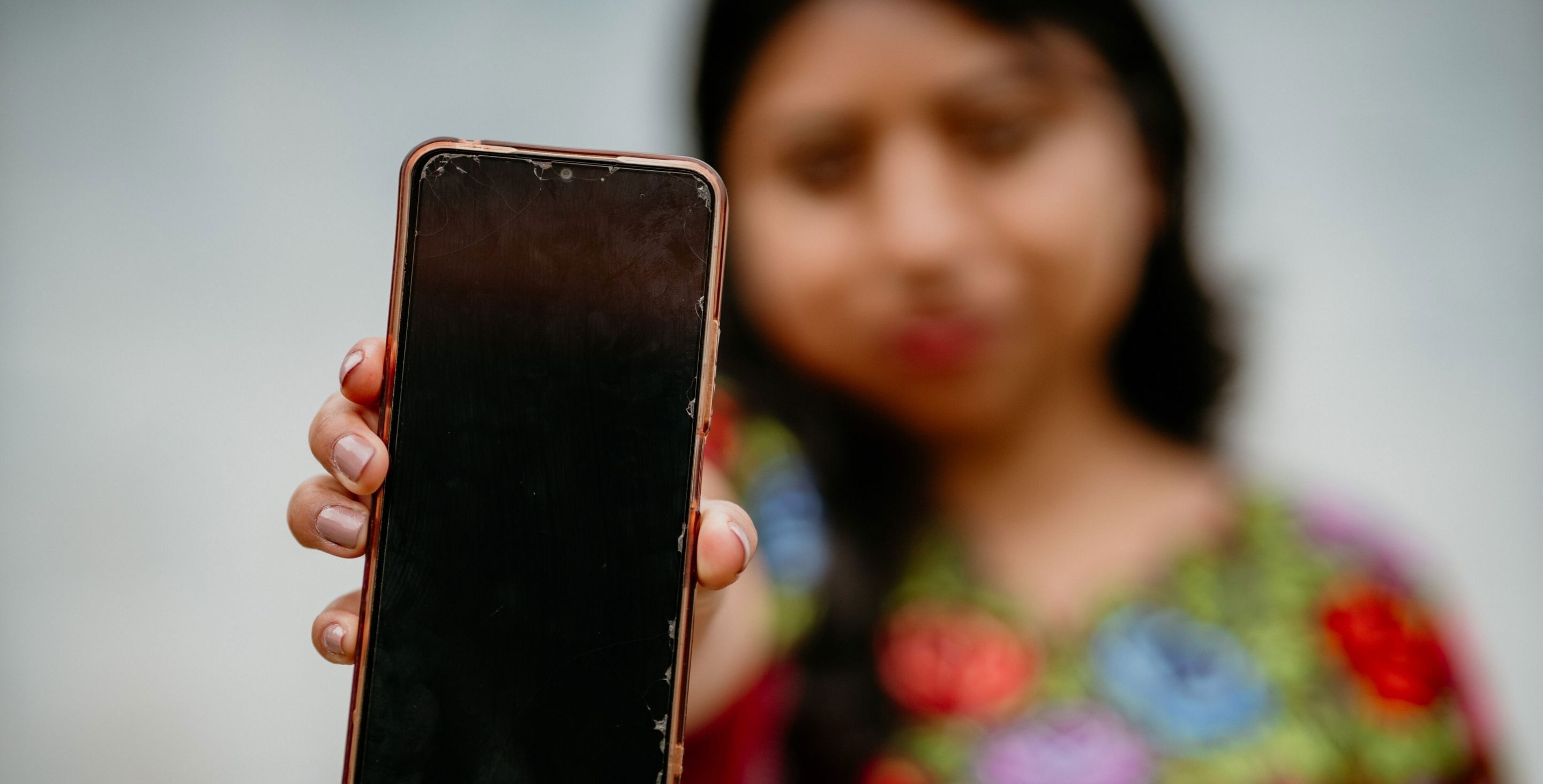 A teenage girl holds a smartphone with a black screen up to the camera.