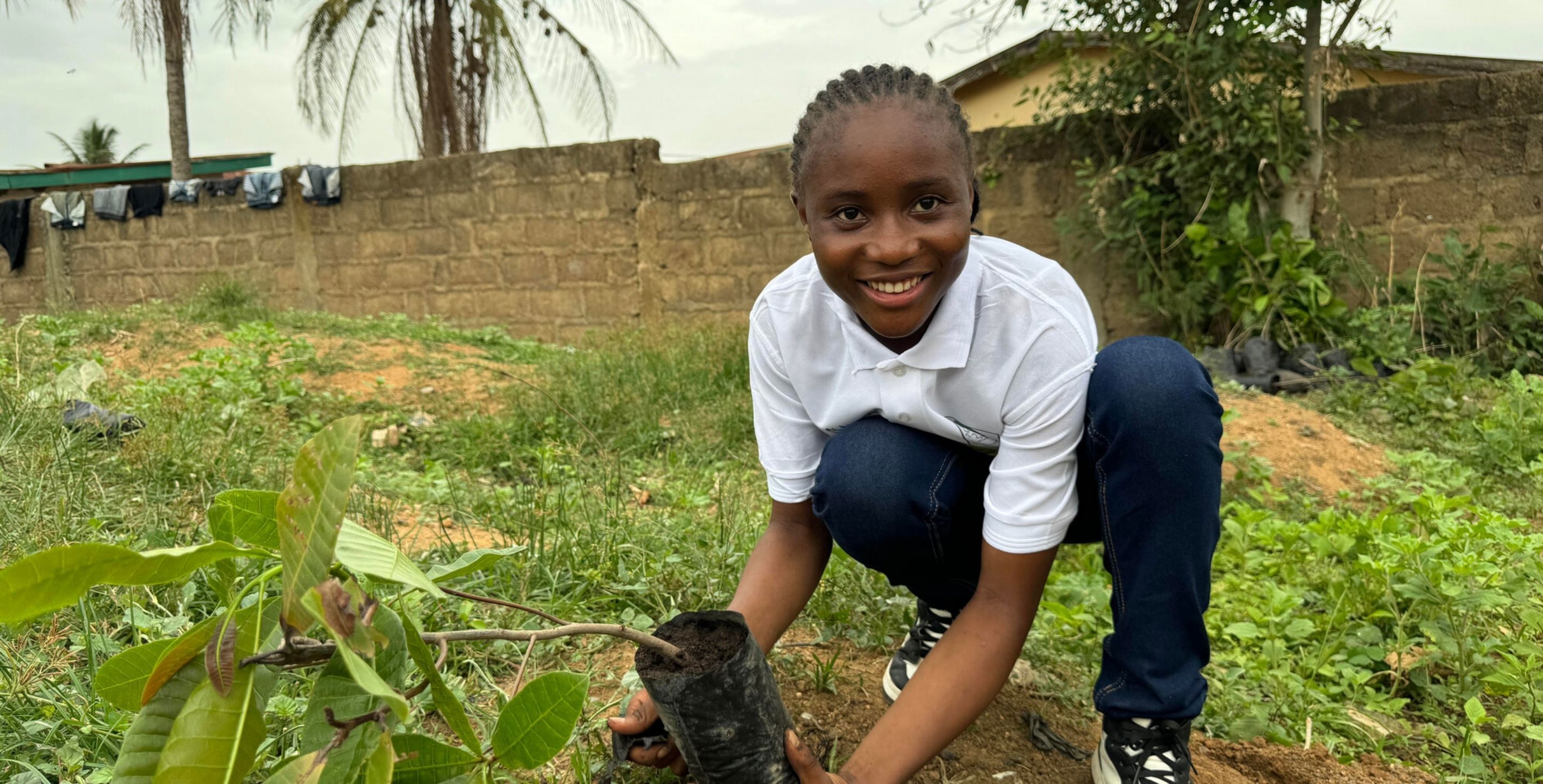 A teenage girl in Sierra Leone plants a tree seedling, smiling at the camera.