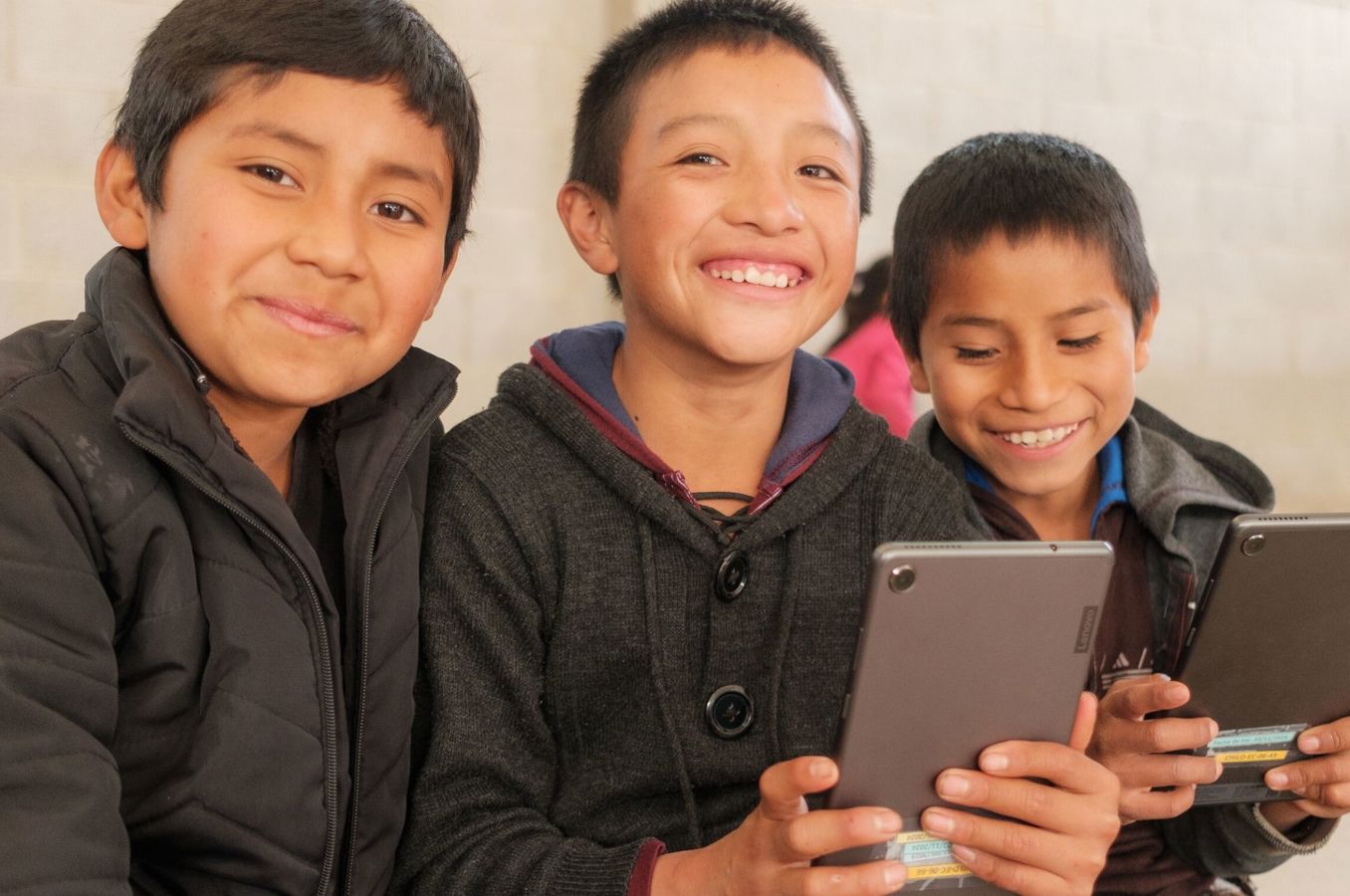 Three boys in Guatemala sit at a desk while looking at cell phones together and smiling.