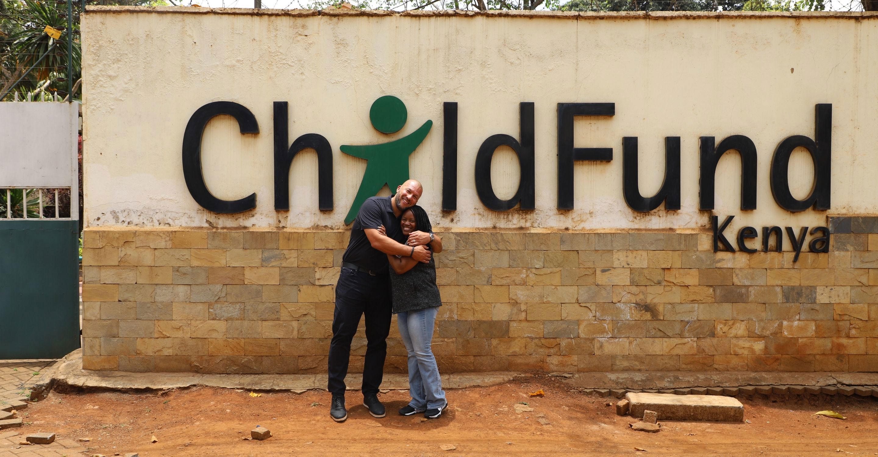 A man and woman stand embracing in front of a large ChildFund Kenya sign.