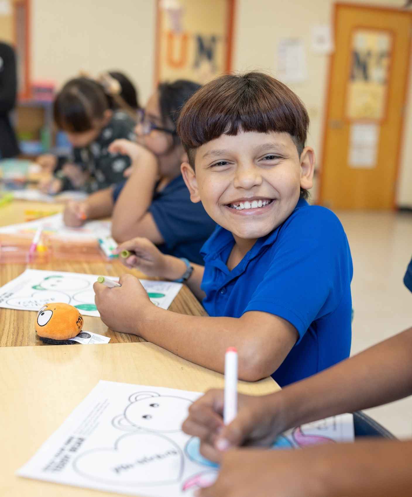 Boy in blue shirt smiling while drawing in a classroom setting.