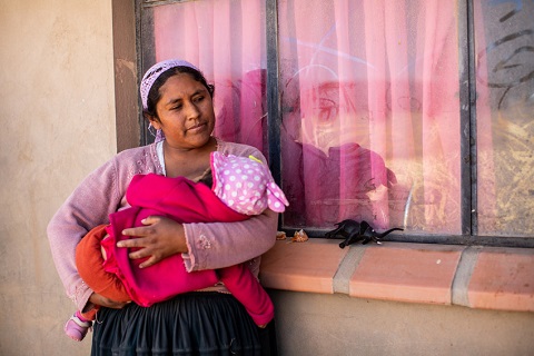 A woman stands outside her home in Bolivia holding a baby in a pink jacket.