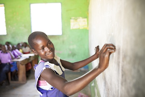 A girl smiles wile she writes on a chalkboard in a classroom in Uganda.