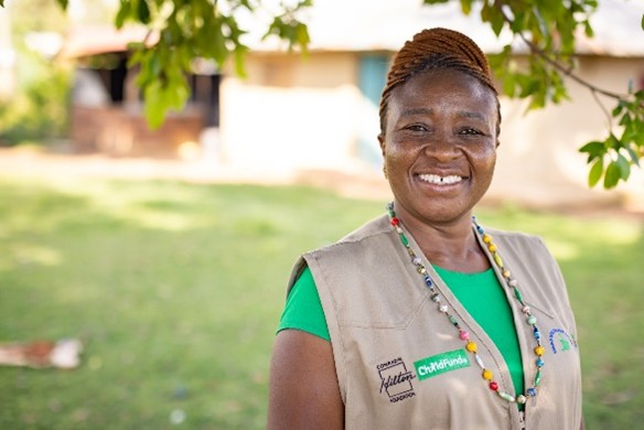 Community health worker standing outside in a rural area, wearing a beige vest with colorful beads and badges.