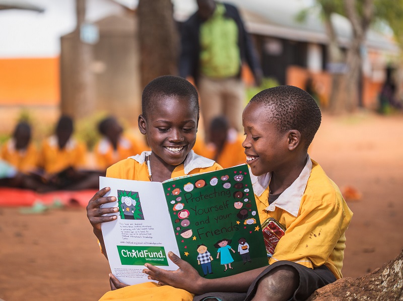Two boys in Uganda share a picture book, laughing, outside.