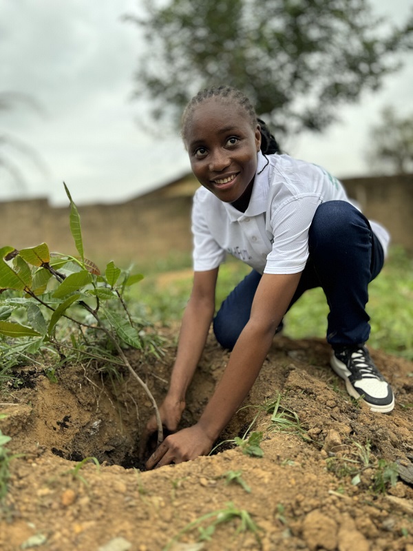 A young girl in Sierra Leone plants a tree, smiling.