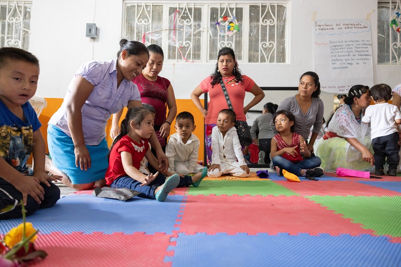 Parents sit in a room with a colorful rug with their preschool aged children.