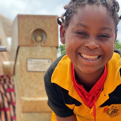 A little girl in Mozambique smiles as she collects water from a borehole into a large bucket.