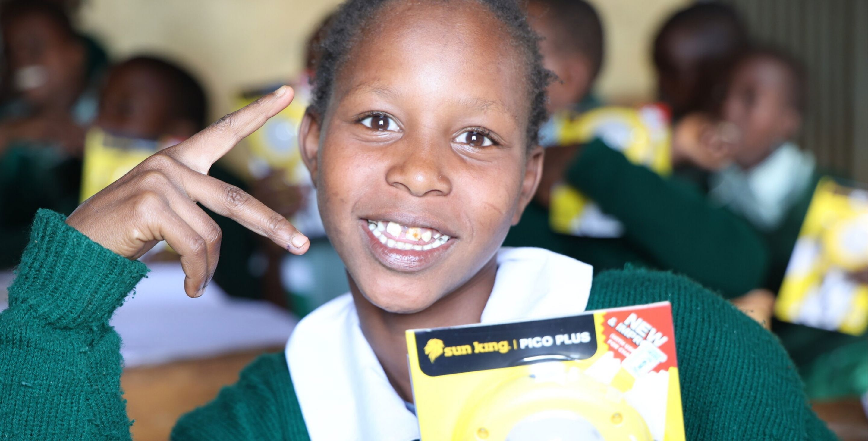 A young girl smiles and flashes a peace sign while holding a solar lamp she received from ChildFund. She is wearing a school uniform and sitting in a classroom.