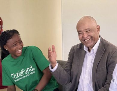 A man in a suit sits with a young woman wearing a green ChildFund T-shirt. Both are laughing.