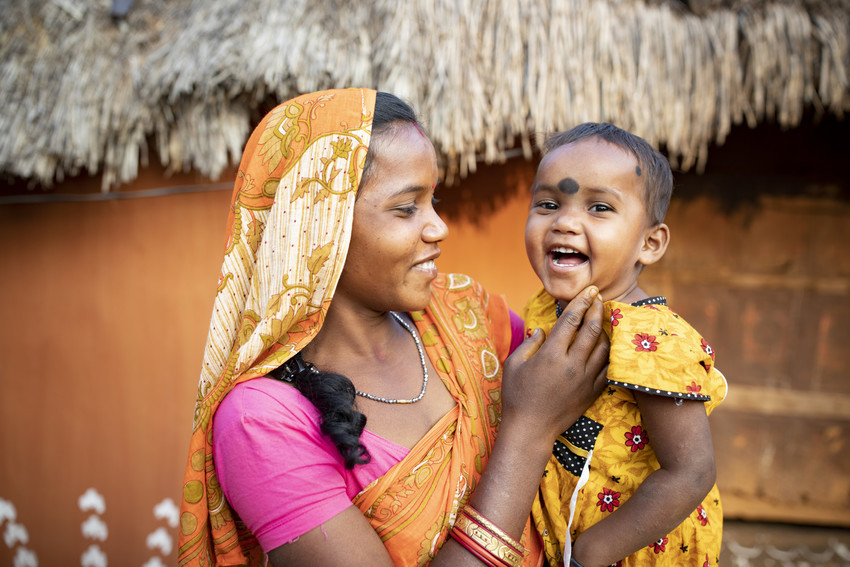 A young mother holds a laughing 2-year-old child in front of a hut in India.