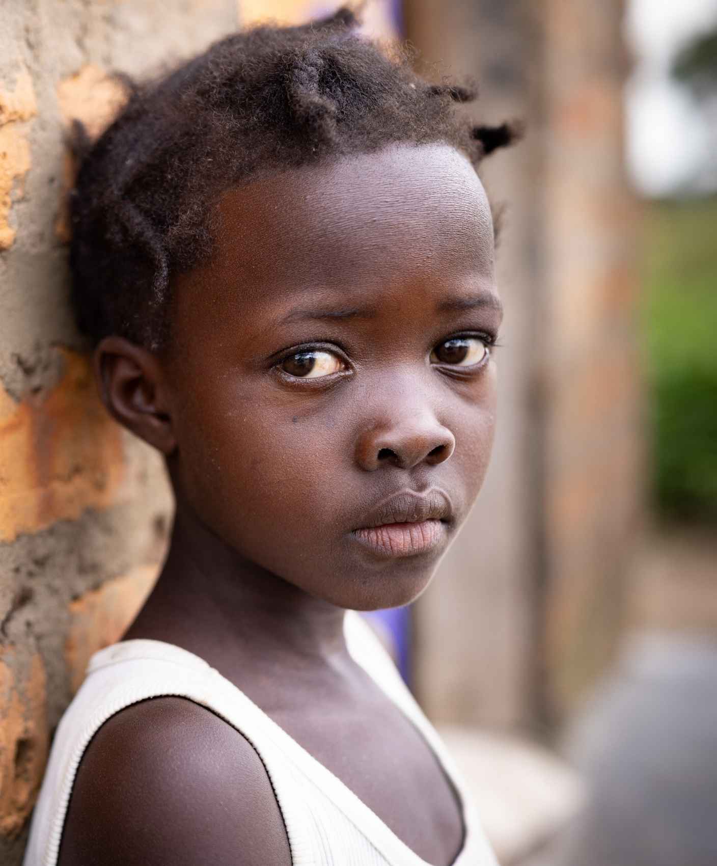 Close-up of a child named Martha leaning against a rough, partially plastered brick wall outdoors in Uganda, wearing a light-colored sleeveless shirt. Martha’s head rests against the wall, facing the camera. The background is softly blurred with earthy ground tones and green vegetation, indicating an outdoor rural setting.