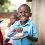 A boy in Uganda wearing a blue school uniform eats from a bowl while smiling widely.