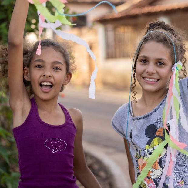 Two nine-year-old girls, Vitoria (in a gray Mickey Mouse shirt) and Ana Clara (in a purple tank top), smile and laugh while playing with colorful streamers outside the House of Culture, a child-friendly community space for socializing and cultural learning.
