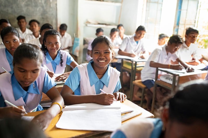 Young girl in India sits at a desk in a classroom, smiling at camera.