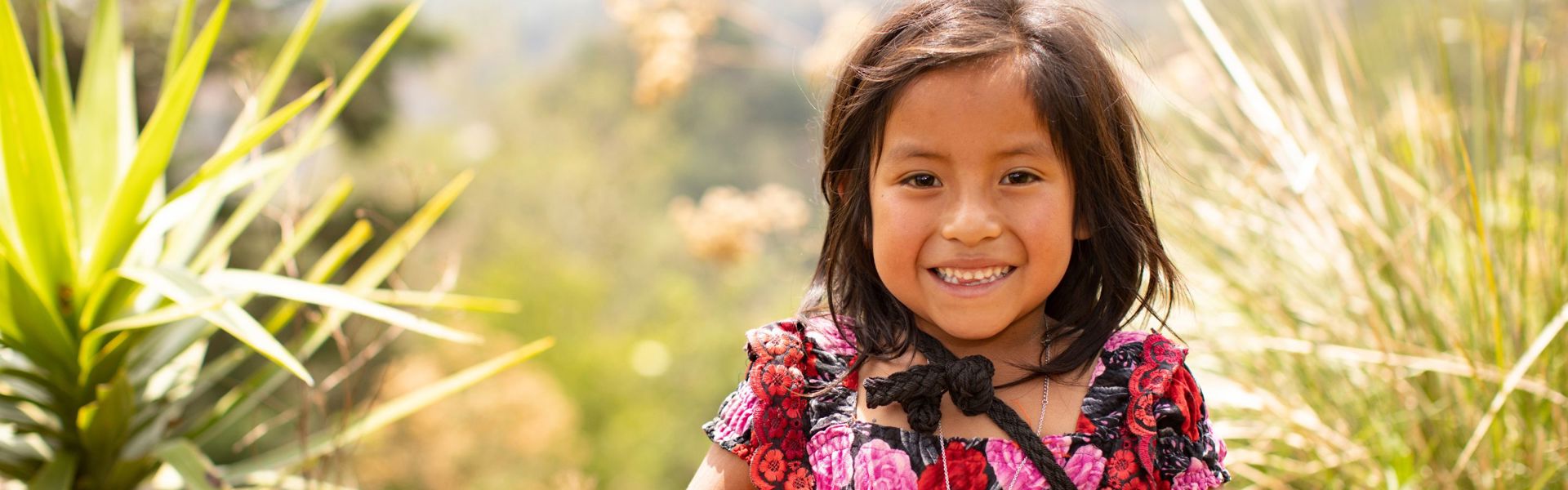 A girl wearing traditional Guatemalan clothing stands outside smiling.