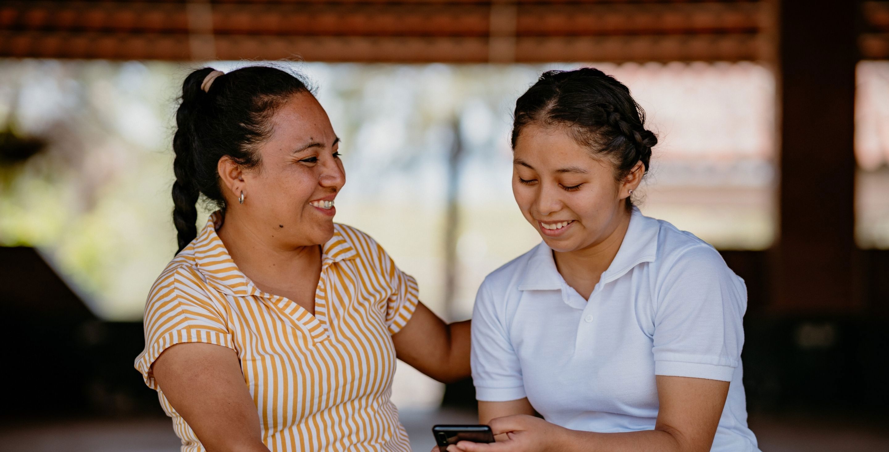 A young woman and an older woman sit outdoors in Guatemala, smiling as they look at a mobile phone together.