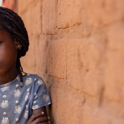 A child in Mozambique sits against the wall of a hut, arms folded, looking pensively at the camera.