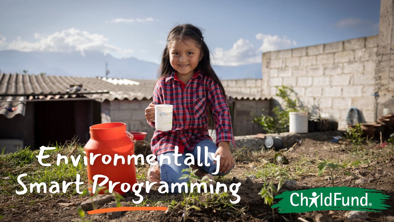 A young girl in a red plaid shirt smiles while kneeling in a garden next to a bright orange water container. She holds a white mug, with mountains and a brick wall in the background. Text overlays read 