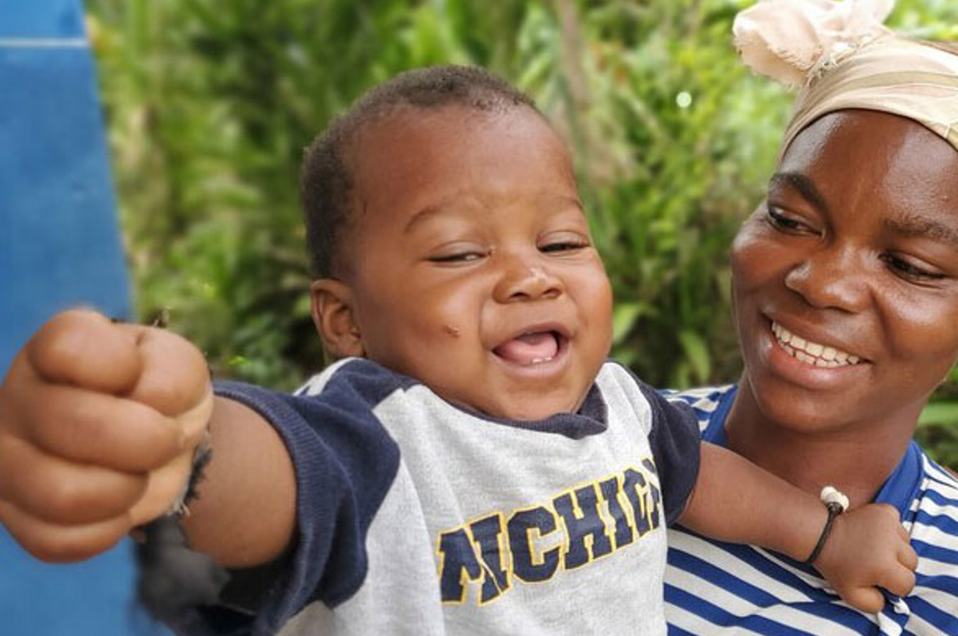 A happy baby in a navy and gray t-shirt reaches out while being held by their smiling mother, who wears a striped shirt and headscarf, with greenery in the background.