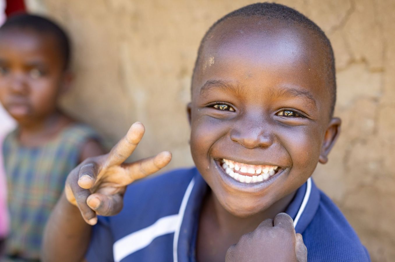 A little boy sitting outdoors in front of an earthen home flashes a peace sign.