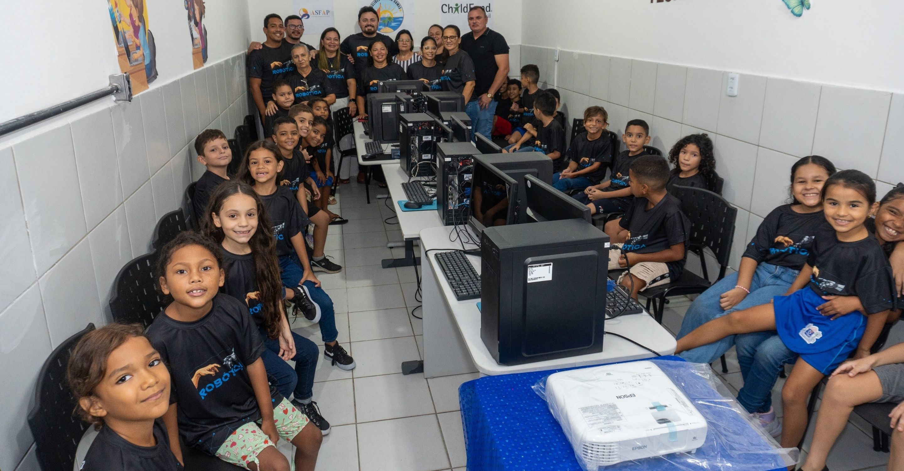 A group of children sit in front of computers in a computer lab, smiling at the camera. 