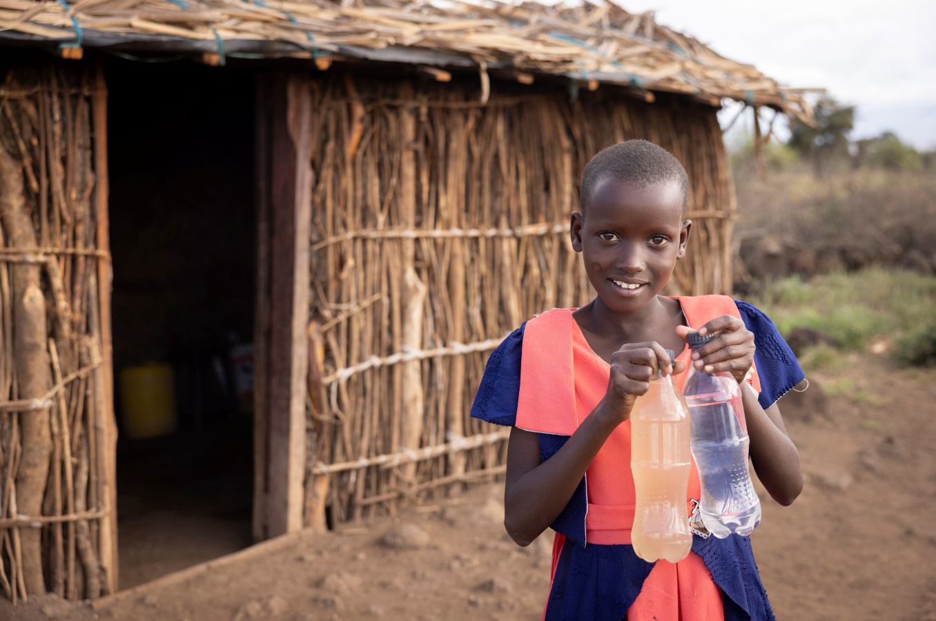 A girl in Kenya standing in front of a hut and wearing a school uniform holds up two plastic bottles of water: one full of murky water, and one full of clean, clear water.