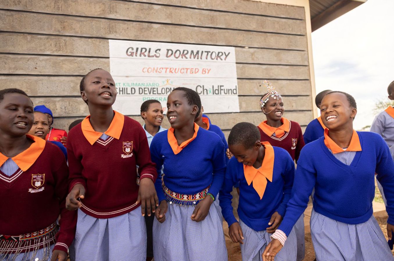 A group of teenage girls wearing school uniforms and traditional Masai jewelry sing and dance together outside a school dormitory.