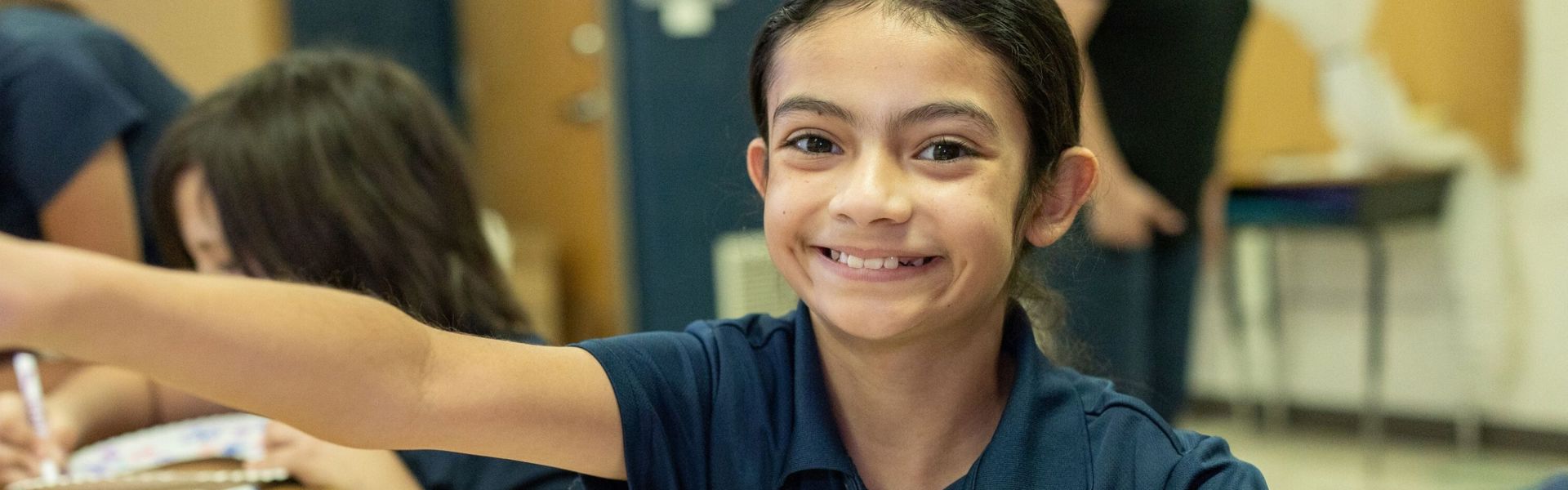 A girl in a U.S. school smiles as she sits at a desk, working on a craft.