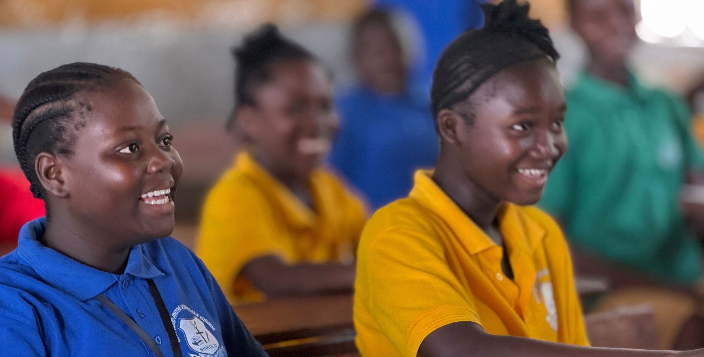 Two teen girls sit in a classroom in Sierra Leone, laughing as they look up at the chalkboard.