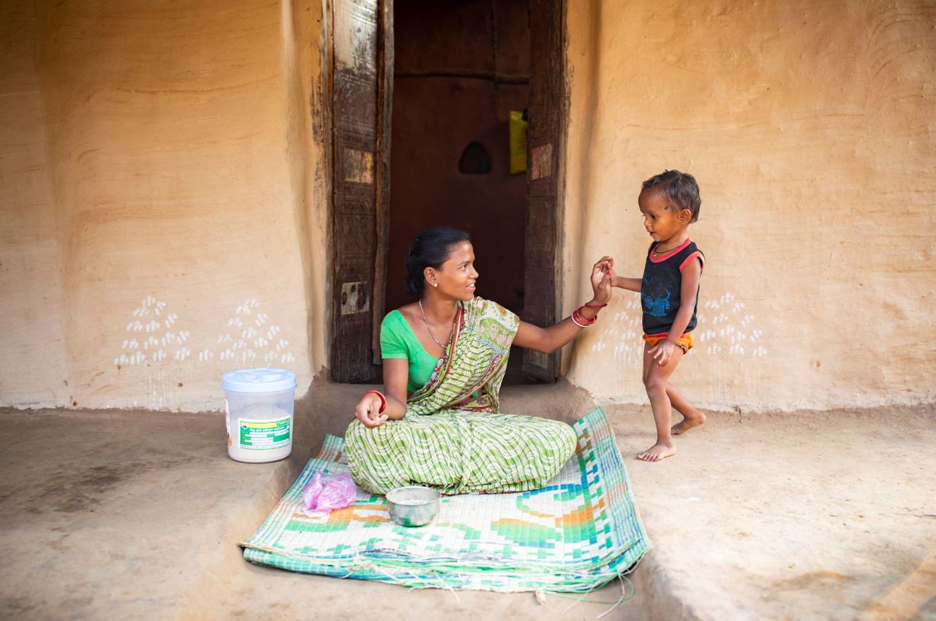 A mom and her toddler sit on their front porch in India, the mom reaching out for the child's hand and smiling. There is a bowl on the mat in front of them and a tub of ChildFund nutritional porridge mix beside them.