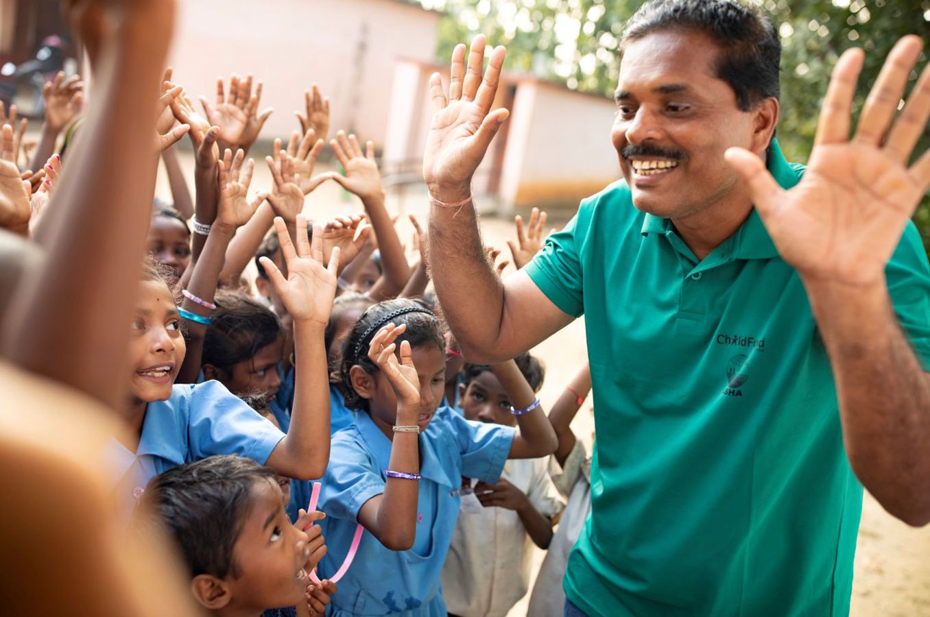 A smiling ChildFund staff member in a green shirt holds up his hands, demonstrating how to properly wash hands. Children crowd around him, smiling and holding up their hands.