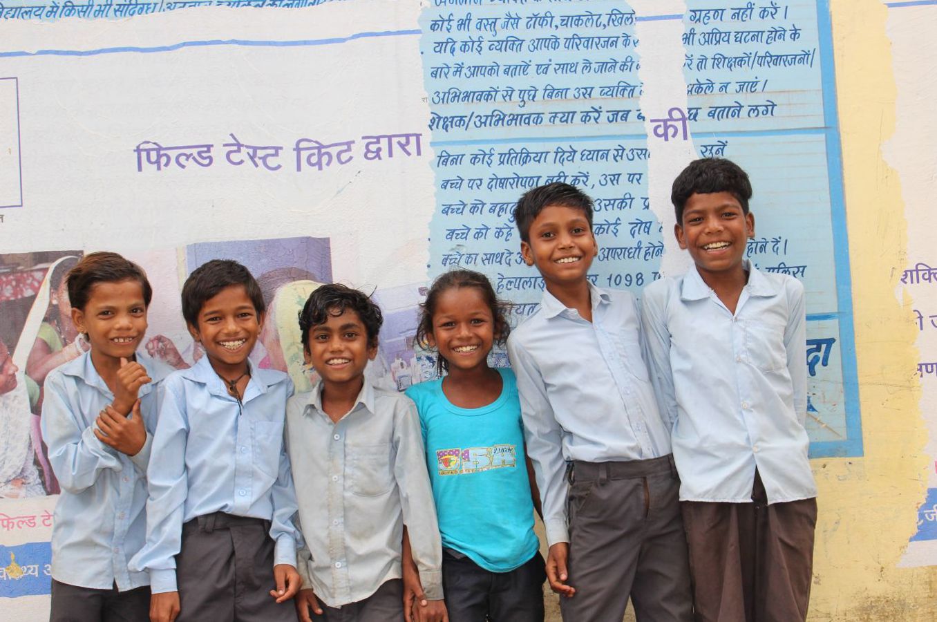 A group of children in India stand in front of the wall of a school inscribed with Hindi lettering.